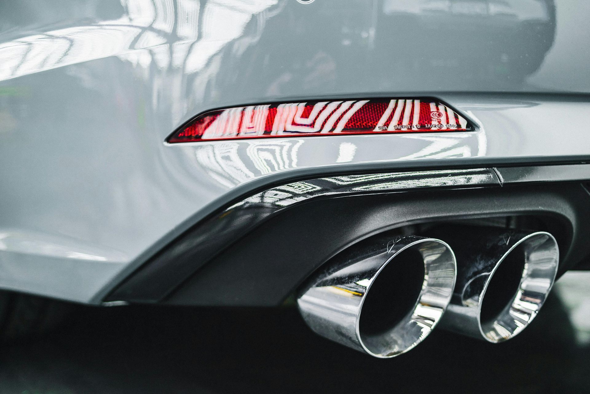 Gray car's rear bumper with chrome dual exhaust pipes and a red reflector.