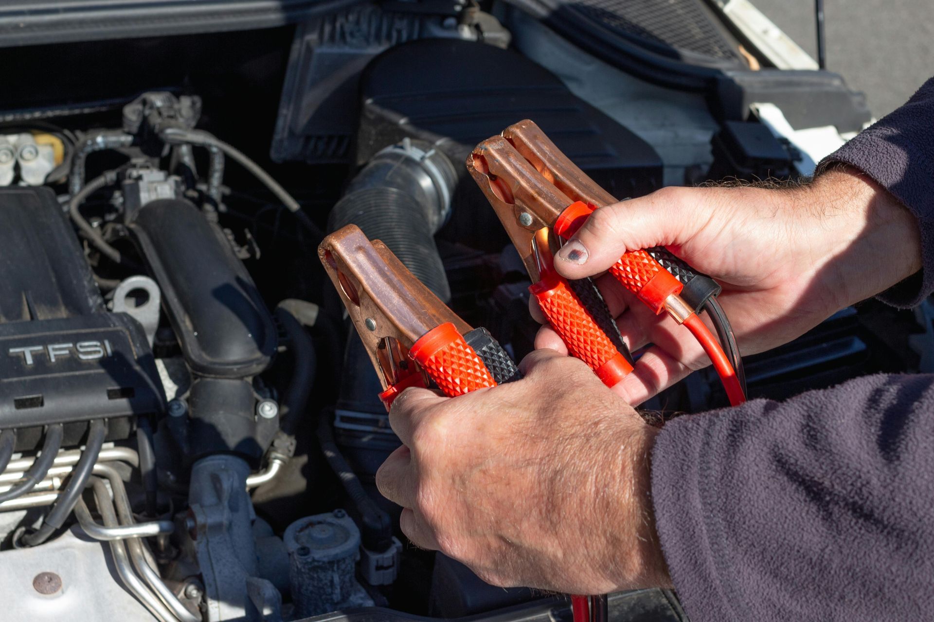 Hands holding red and black jumper cables, connecting to a car engine.