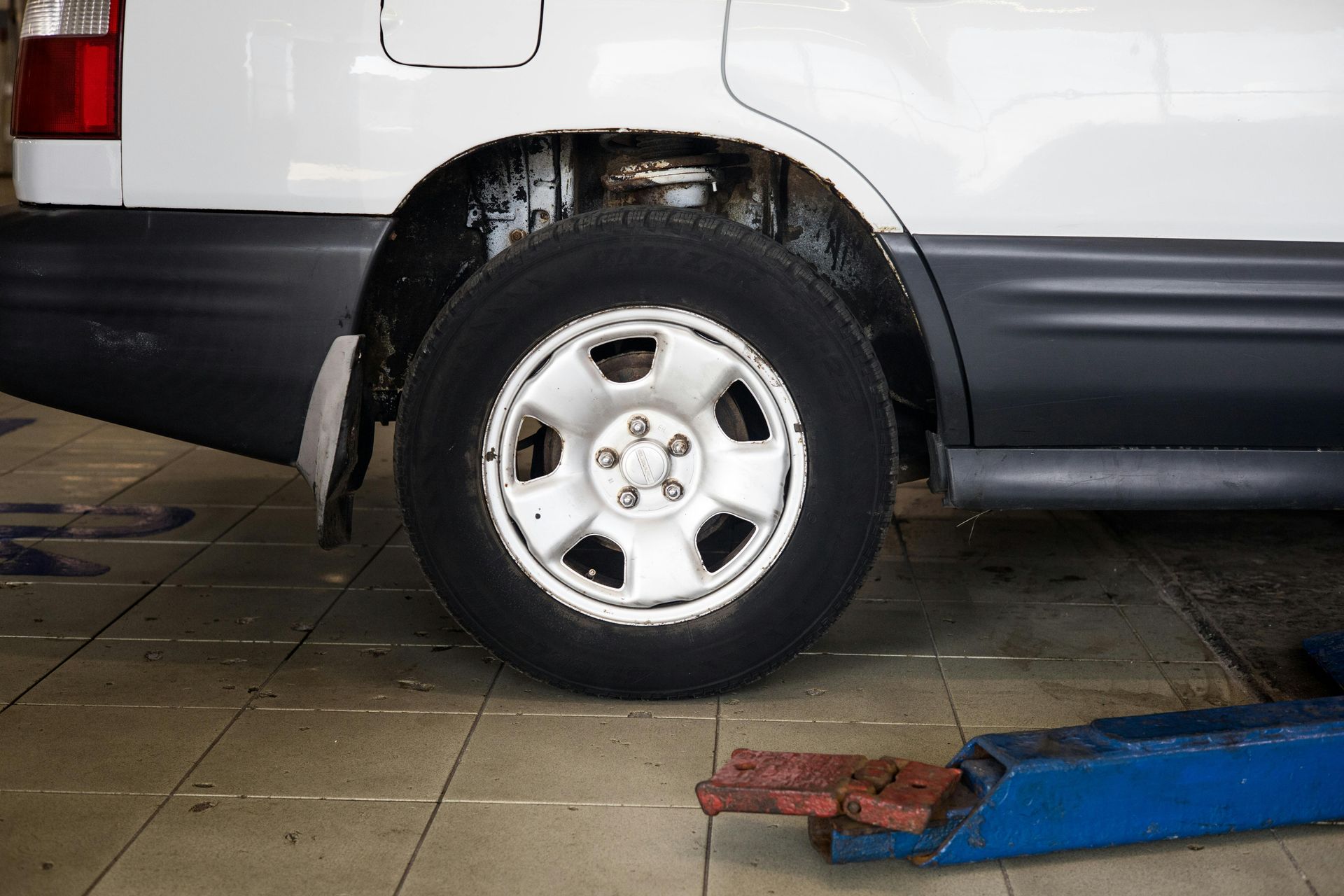 White car's rear tire in a garage, visible suspension. The vehicle sits on a tiled floor with a car lift nearby.