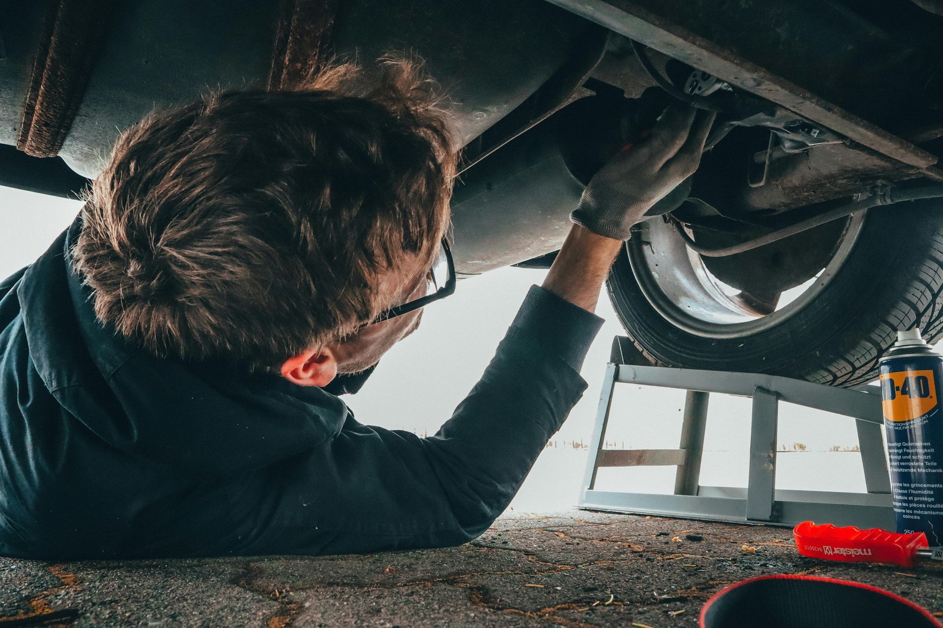 Person working on the underside of a car, using tools. Kneeling on the ground, next to a car wheel.