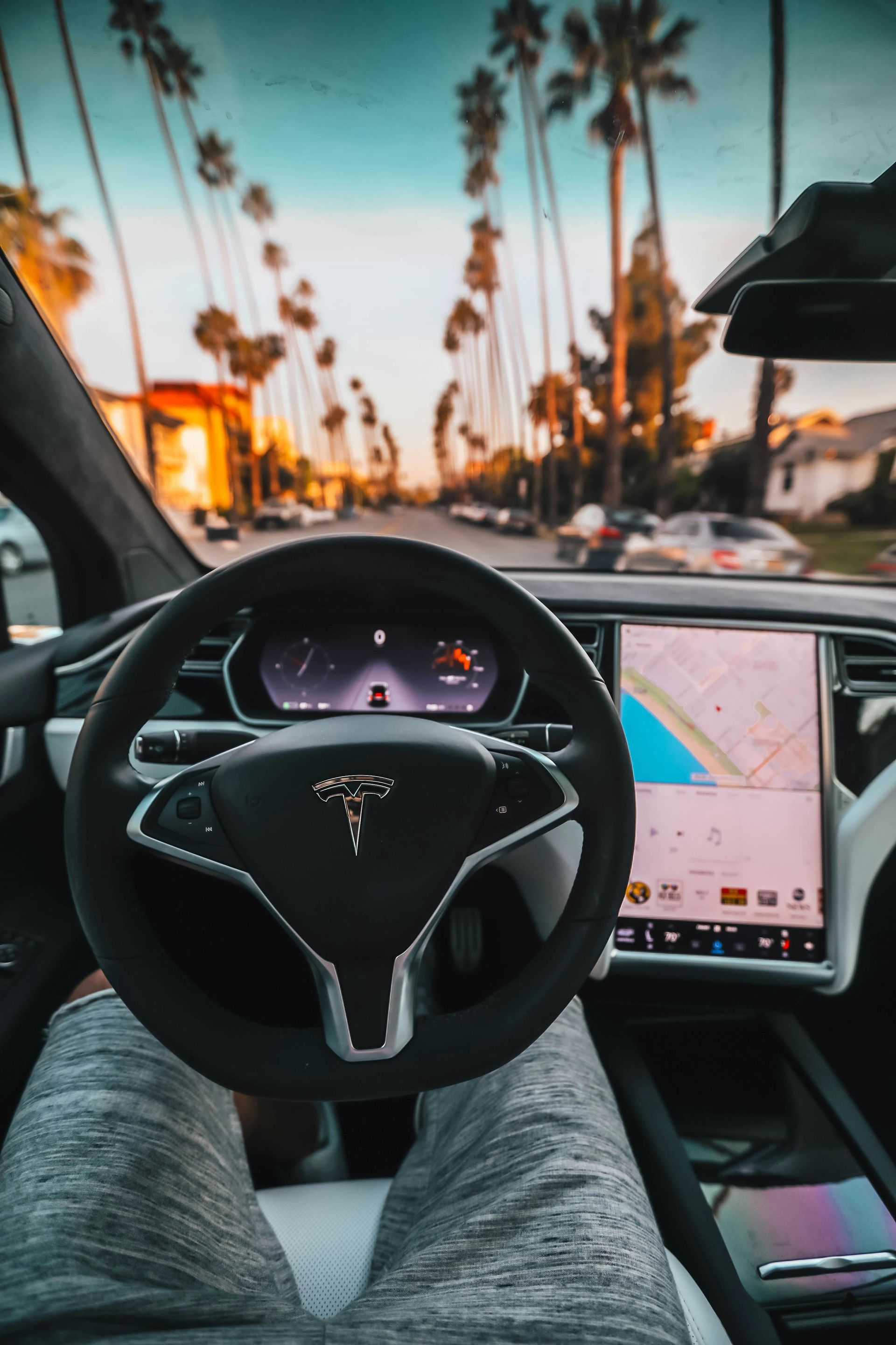 A first-person view from the driver's seat of a Tesla, looking down a palm tree-lined street at sunset.
